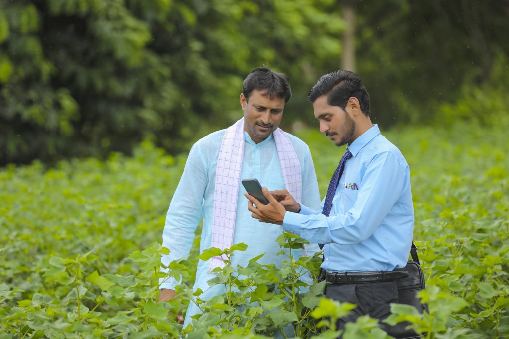 young indian agronomist or banker showing some information to farmer in smartphone at agriculture field.