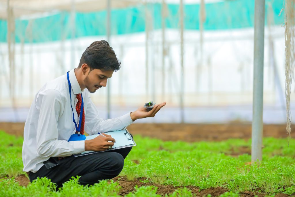 young indian agronomist holding small plant in hand and collecting some information at greenhouse